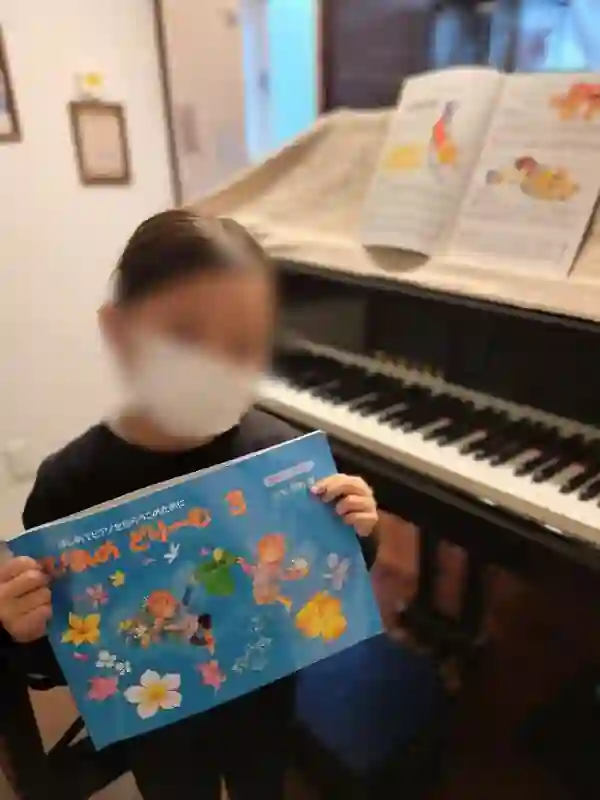 N piano music schoolのレッスンで「ぴあのどりーむ3」の教材を持つ幼い女の子。グランドピアノの前で学びの記録として記念撮影。