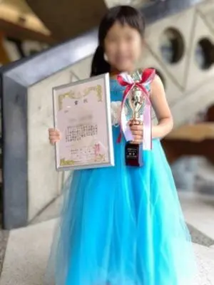 N piano music schoolの生徒が、ピアノコンクールで金賞を受賞し、トロフィーと賞状を手に記念撮影。ブルーのドレスが印象的な晴れ姿。