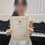 小山市のピアノ教室N piano music schoolの中学生の生徒がブルグミュラーコンクールで優秀賞を受賞、ピアノの前で賞状を持ち笑顔の写真